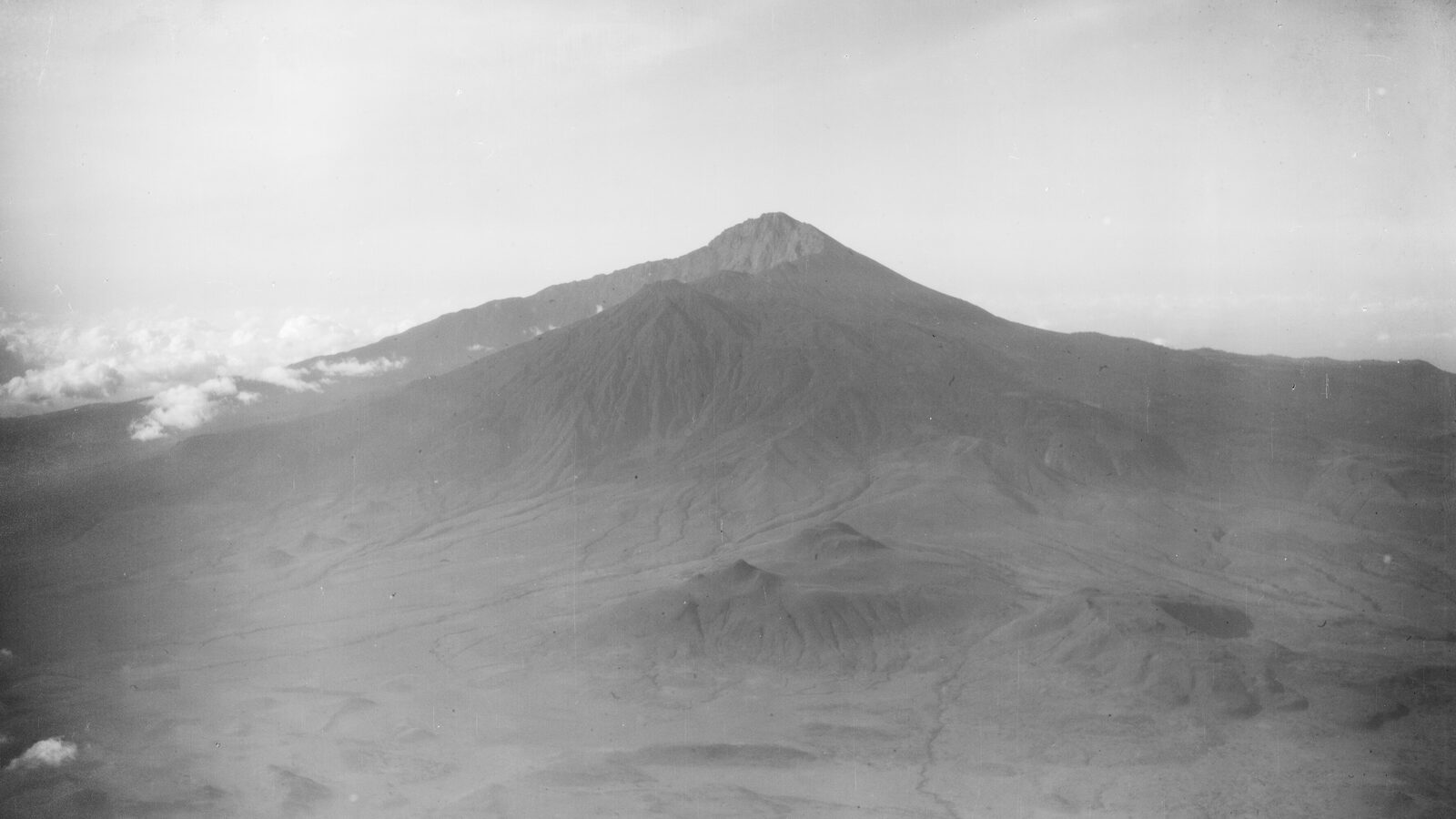 Aerial photograph of Mount Meru in northern Tanganyika, its summit crater breached to the east, rising above a plain scattered with smaller volcanic cones.